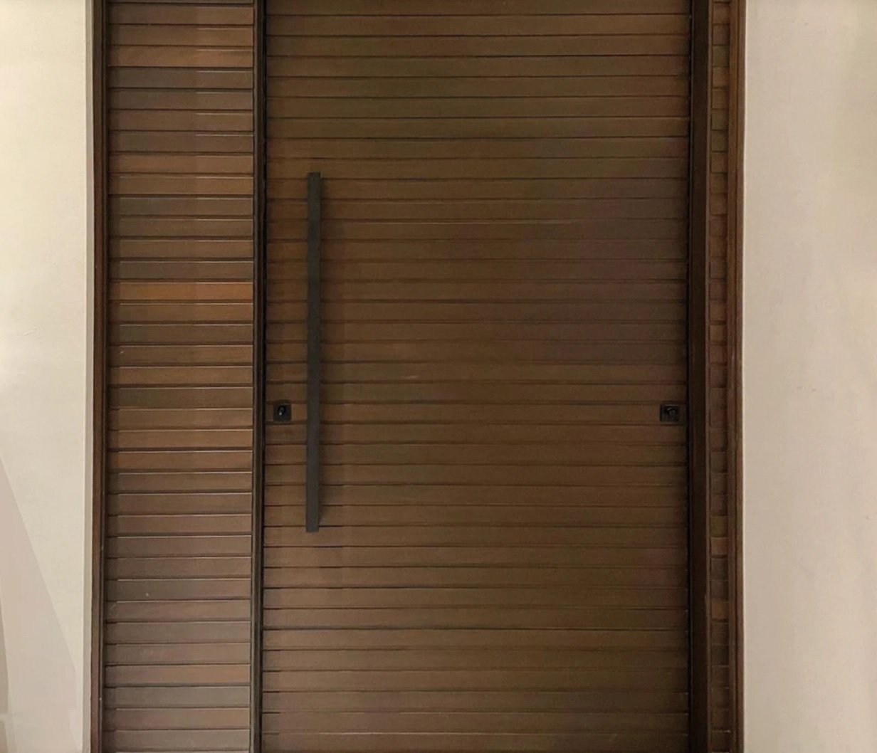 Dark hardwood pivot door made of horizontal slats with a matching slatted side panel and a square bar pull